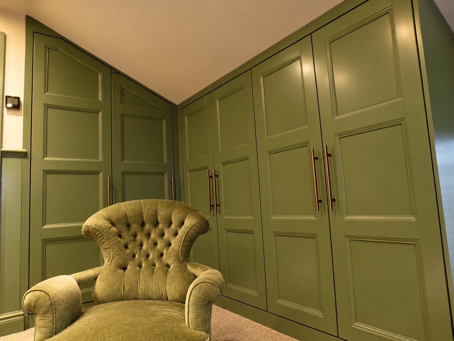 Green panelled wardrobes fitted beneath the sloping bedroom ceiling beside a buttoned armchair